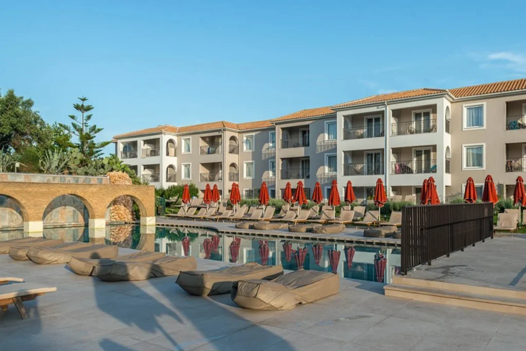 Spacious outdoor swimming pool surrounded by sun loungers, red parasols and contemporary hotel buildings at Bitzaro Grande Hotel & Suites, Zakynthos.