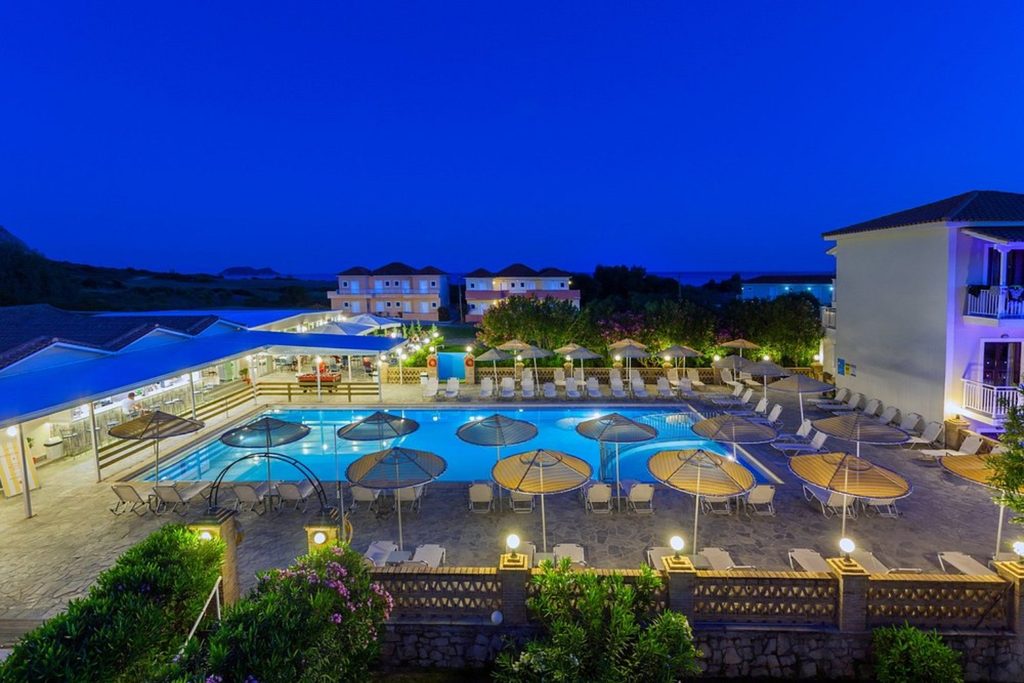 Illuminated outdoor swimming pool with sun loungers and parasols in the evening at Cavo D’Oro Hotel, Zakynthos
