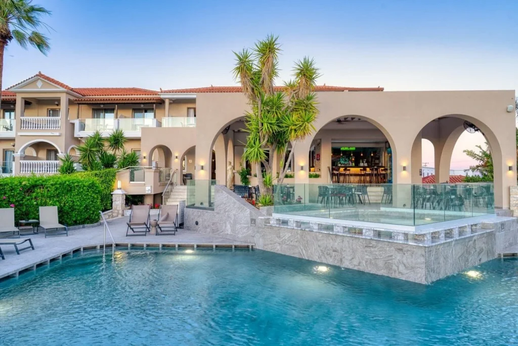 Modern outdoor swimming pool surrounded by sun loungers, tropical plants and an arched poolside bar and terrace at Venus Hotel & Suites, Zakynthos.